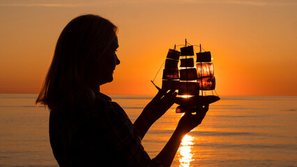 Silhouette of a young woman with a toy boat in her hands. Stands against the backdrop of the sea where the sun sets