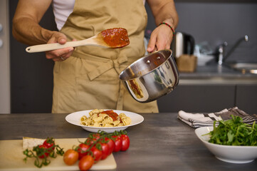 Close-up chef pouring tomato sauce on Italian pasta, plating up the dish before serving. Man cooking dinner at home kitchen. Cuisine. Culinary. Epicure. Food concept