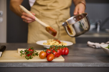A branch of fresh ripe organic tomato cherry on the cutting board against the backdrop of a blurred male chef cooking Italian pasta at home kitchen. Traditional Italian cuisine. Food background