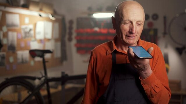 Bicycle mechanic old man working in a garage repairing bicycles stands dictating a voice message and using a virtual assistant on a smartphone