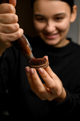 Focus on half of macaroon on which pretty smiling female confectioner applies chocolate cream