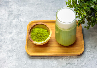 Matcha latte green tea with milk foam in tall glass  with a small bowl of matcha green tea powder in wooden bamboo plate isolated on grey background, copy space.