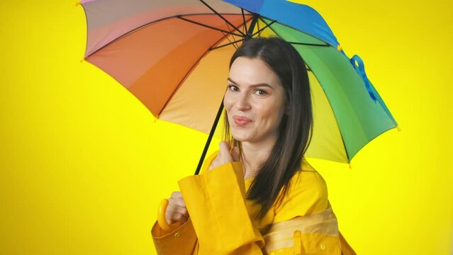 A happy attractive woman in a bright yellow raincoat with a rainbow umbrella is checking for the rain. Yellow studio background
