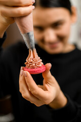 Close-up focus on red half of macaroon on which female pastry chef applies pink cream