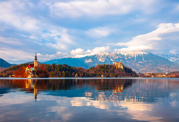 The beautiful surroundings of Lake Bled in Slovenia with Bled island and its little chapel in the foreground, reflected in the calm waters of the lake. Flanked by the fall colored forest and mountains