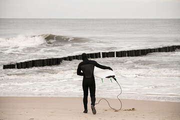 Slender male surfer in a black wetsuit walks into the water, holding a surfboard in his hand