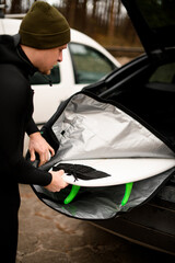 Young male surfer takes out a surfboard from a cover lying in the trunk of his car