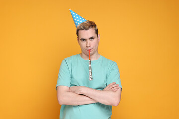Sad young man with party hat and blower on orange background