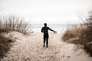 Young male surfer, dressed in a wetsuit, walks on the sand between the bushes