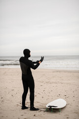 Male surfer standing on the seashore next to his surfboard and wearing surfing gloves