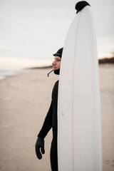 Surfer man in black wetsuit standing on sandy beach and looking thoughtfully at sea