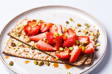 Matza toast with peanut butter, strawberries and pistachios on a white plate. Traditional bread for the Jewish holiday of Passover. - obrazy, fototapety, plakaty