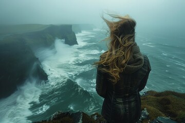 A woman in contemplative stance observes the foggy ocean cliffs wearing a snug sweater and a scarf