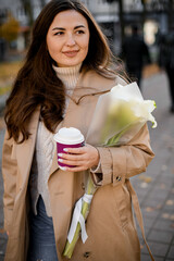 Cute smiling woman standing outdoors holding a cup of coffee and a bouquet of white lilies
