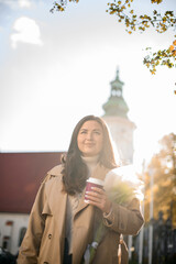 Cute smiling woman standing outdoors holding a cup of coffee and a bouquet of white lilies illuminated by sunbeams