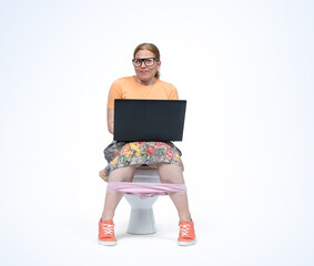 A happy young woman in glasses and a colorful skirt is sitting on the toilet with a laptop on her lap, isolated on a light blue background