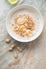 Bowl with peanut butter and banana oatmeal, vertical shot on a beige and roseate granite background, flat lay with space