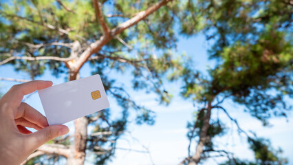 A white bank card is held in a woman's hand, set against the backdrop of a pine tree and the sky.