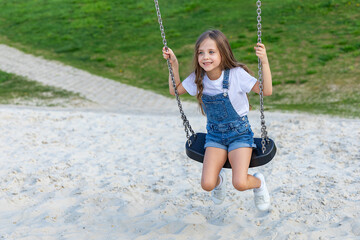 a happy little girl rides on a swing on a playground in summer on a sunny day. Children's Day