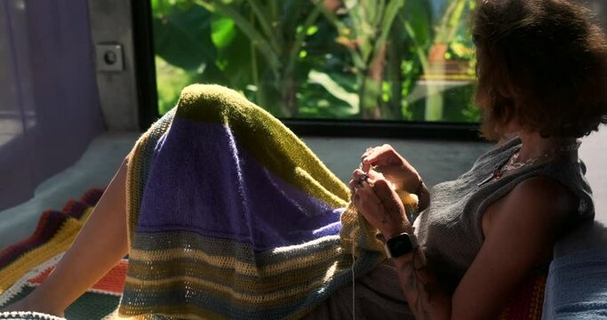A woman sits on the floor in the bedroom and knits a blanket from woolen threads. The background in the window is tropical greenery. Next to her is a basket with balls of wool and knitting tools.