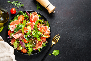 Green salad on black background. Fresh salad with jamon, green salad leaves and tomatoes. Top view with space for text.