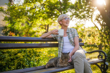 Happy senior woman enjoys reading book and spending time with her cat while sitting on a bench in her garden. - 790819701