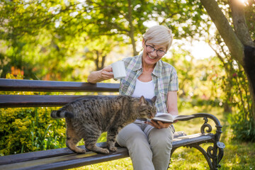 Happy senior woman enjoys reading book ,drinking coffee  and spending time with her cat on a bench in  garden. - 790819723
