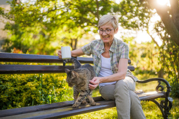Happy senior woman enjoys drinking coffee and spending time with her cat while sitting on a bench in her garden. - 790819732
