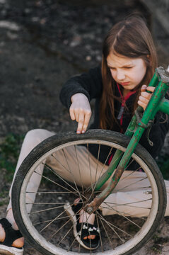 Little sad teenage girl, dissatisfied distressed child sitting near an old bicycle with a broken, punctured wheel tire outdoors. Photography, portrait.