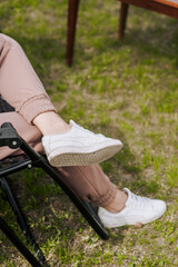 Young woman relaxing on vacation, sitting on a chair on green grass outdoors. Close-up photo of feet with sneakers.