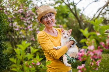 Portrait of happy senior woman holding her cute cat while gardening in yard.