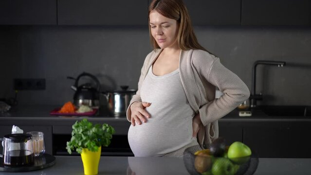A pregnant woman is thinking about what to eat, while standing in a modern kitchen in her apartment. A woman takes a green apple from a fruit bowl, that is on the table and smiles