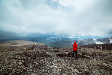 Man in red jacket on high stony pass with view to valley against large snow-capped mountain range in rainy low clouds. Guy among sharp stones on hill. Misty snowy mountains in rain under cloudy sky. - 790836160