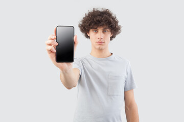 Handsome young man showing the blank screen of his smartphone to the camera, while looking into the camera with his blue eyes, isolated against a gray background