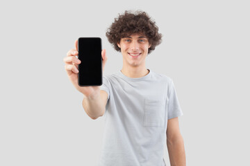 Smiling and handsome, the young man showing the blank screen of his smartphone to the camera, while looking into the camera with his blue eyes, isolated against a gray background