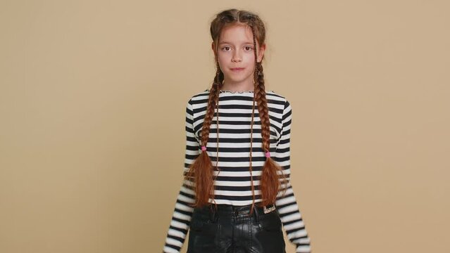 Cheerful funny young school girl showing tongue making faces at camera, fooling around, joking, aping with silly face teasing bullying, abuse. Preteen female child kid 10 years old on beige background