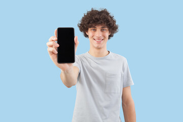 Smiling and handsome, the young man showing the blank screen of his smartphone to the camera, while looking into the camera with his blue eyes, isolated against a blue background