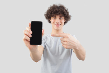 Smiling and handsome, the young man showing the blank screen of his smartphone to the camera pointing display with finger while looking into the camera with his blue eyes, isolated on gray background