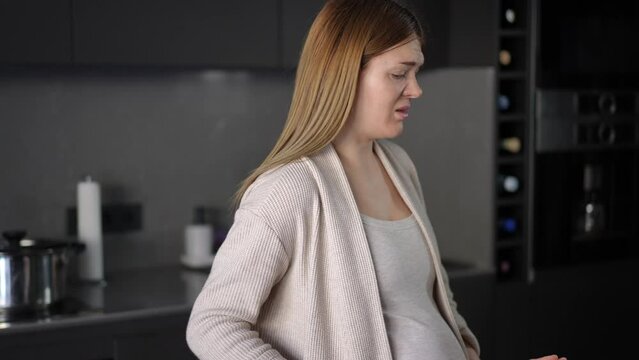 Close-up. A pregnant woman sniffs an onion and feels intense disgust, grimacing and waving her arms while standing near the table in the kitchen. Odors during pregnancy