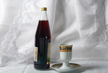 Bottle is dark with a golden cap of grape juice and a Kiddush cup against a white lacy curtain. The cup has colorful designs and sits on a matching saucer. Shabbat, passover, pesah celebration concept