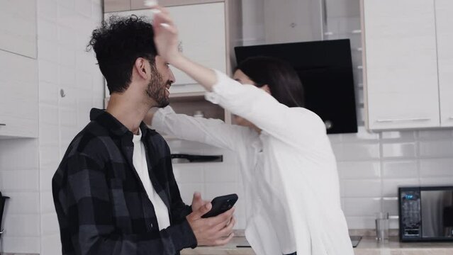 Young woman looking at the smartphone of her partner, becoming surprised and overjoyed, and they are starting to hug together in cozy kitchen at home