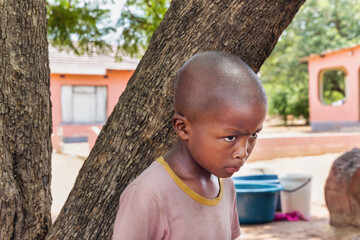 abused upset village african child, hiding behind a tree, standing in front of the house,