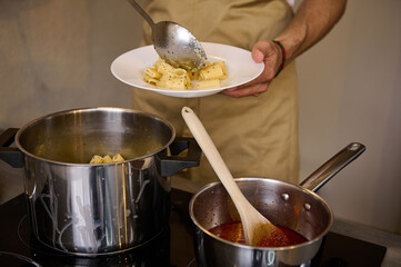 Close-up male chef plating up freshly cooked pasta, putting boiled penne on a white plate, standing at induction electric stove with saucepan full of tomato pasta. Italian culinary. Food background
