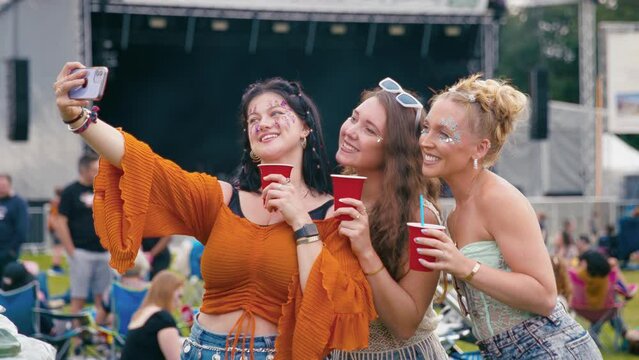 Three female friends wearing glitter and holding drinks posing for selfie at outdoor summer music festival - shot in slow motion 