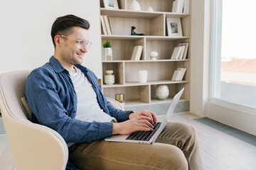 Contented man works on a laptop, comfortably seated in an elegant chair by the bookshelf in a...