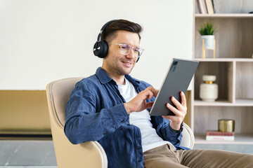 Man in casual attire enjoys digital content on a tablet while wearing headphones, seated in a...