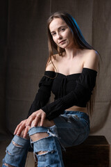 Classic studio portrait of a young brunette dressed in a black top, who is sitting on a chair against a black background.