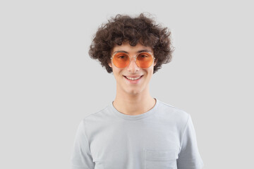 Smiling and handsome, the young man wears sunglasses. He looks into the camera with his eyes, a portrait of a man with orange lens eyewear isolated against a gray background