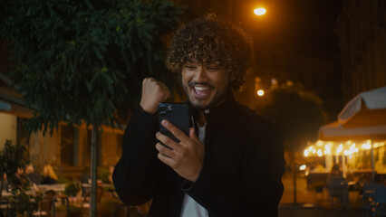 Indian Arabian Latino American man holding smartphone mobile phone cellphone telephone looking at screen rejoice victory good news luck joy success celebrate win amazing screaming excitement male guy