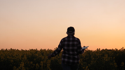 Farmer with digital tablet walking away to rapeseed field. Agriculture business. Harvesting of agricultural products. Silhouette of woman agronomist at sunset light in rural field bloom canola crop